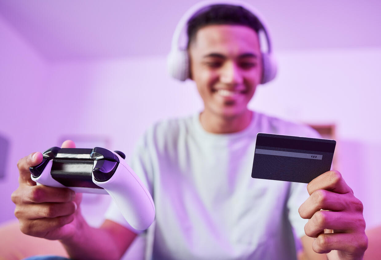 teen boy with headphones, holding a video game controler in one hand and a credit card in the other