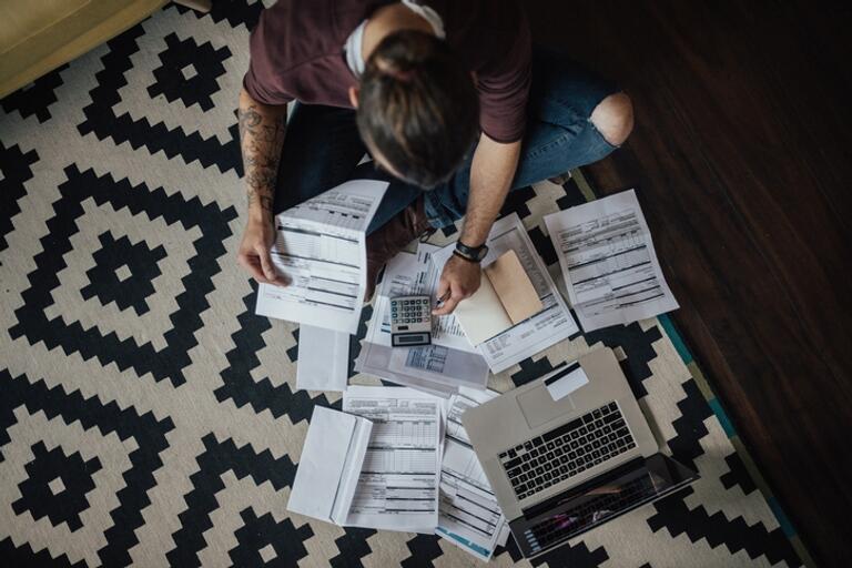 person sitting on the ground with bills, and other papers with a laptop open. 