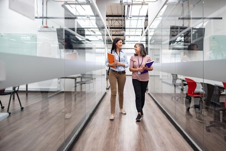 two women walking down a long hall, that has glass walls. 