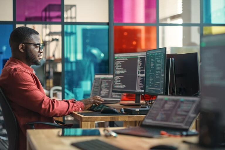 man on computer with several screens and a large colorful glass window behind.