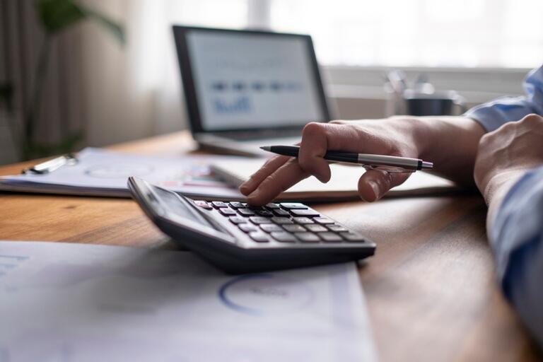 desk with someone using a calculator, with a laptop open on the desk behind, and papers scattered on the desk.