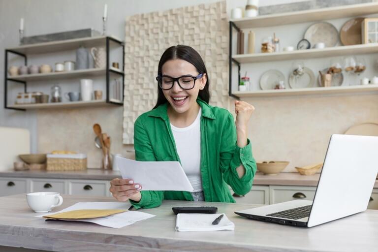 woman in a kitchen setting looking cheerful at a peice of paper, her laptop is open next to her.