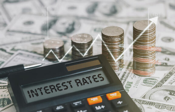 calculator with the words interest rates on it with change stacked up in the background. Money and a line chart are also in the background.