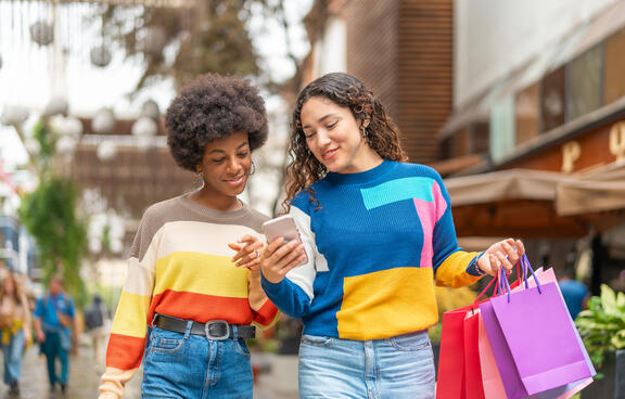 two girls wearing bright sweaters, one is holding a phone and lots of shopping bags.