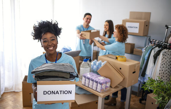 Group of tees volunteering, filling boxes marked "donations" with a young black woman in the front of frame holding a full box of donated items.